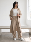 A woman stands in a bright, minimalist room wearing a beige long open-front kimono over a white top and matching beige wide-leg drawstring pants, paired with white sneakers.