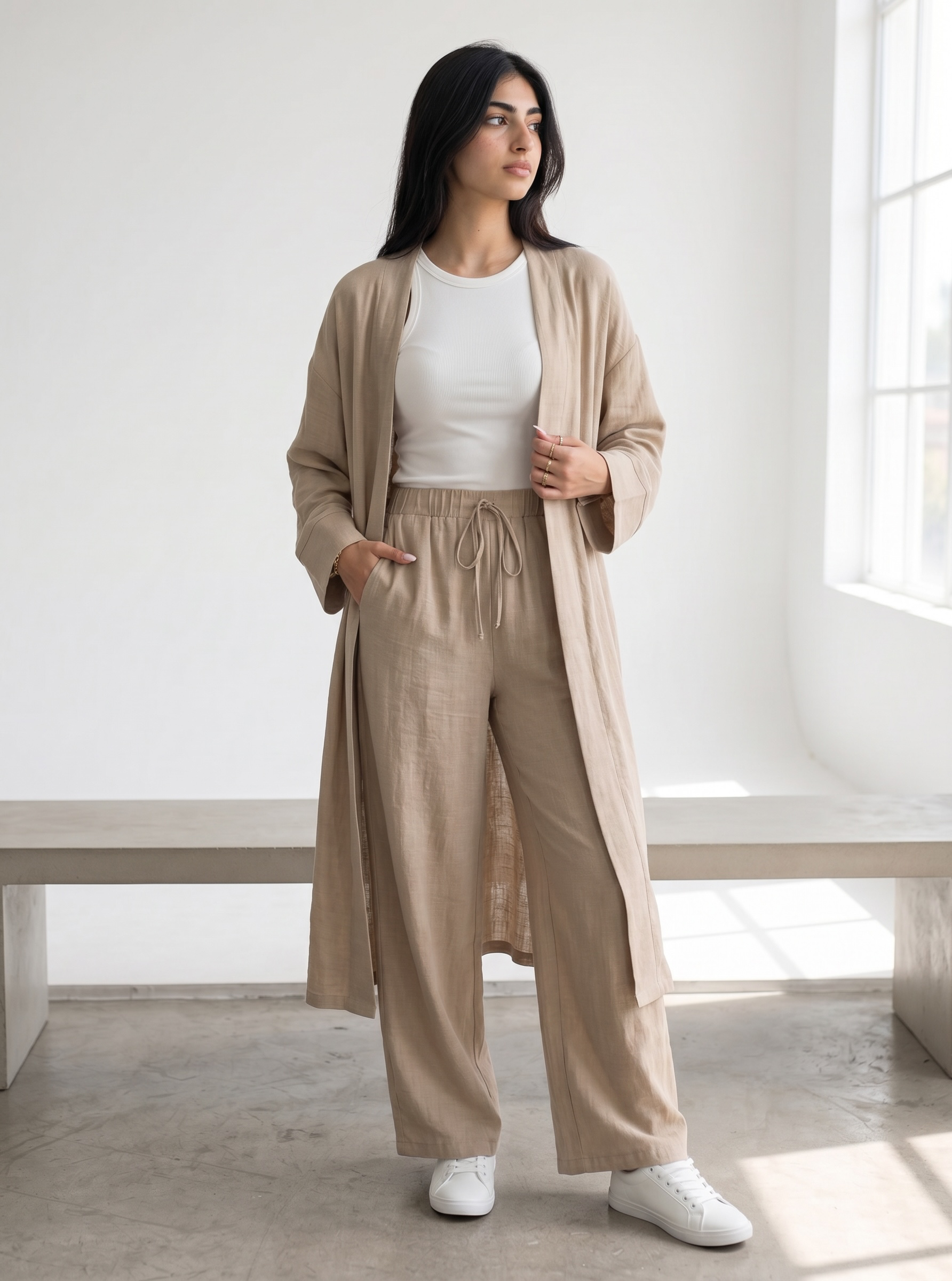 A woman stands in a bright, minimalist room wearing a beige long open-front kimono over a white top and matching beige wide-leg drawstring pants, paired with white sneakers.