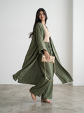 A woman modeling a matching olive green two-piece outfit consisting of a long-sleeve open-front cardigan and wide-leg pants, paired with a beige crop top, beige sandals, and a beige clutch bag.